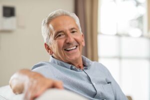 a man sits on a couch smiling, showing off his hybrid dentures.